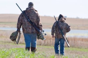 Father and son wearing jeans and camo jackets have their shotguns and hunting gear as they walk onto a field