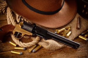 A handgun resting on a surface, surrounded by bullets. The background has a cowboy hat, burlap, and a rope
