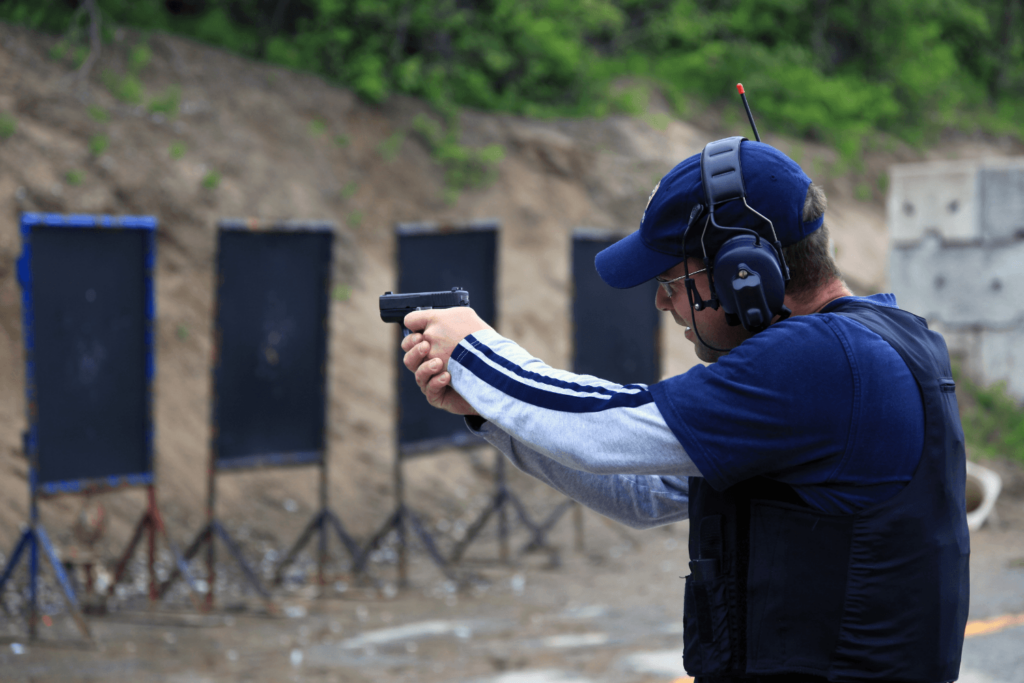Man at an outdoor shooting range wearing dark clothes and protective gear aims his pistol at a target 