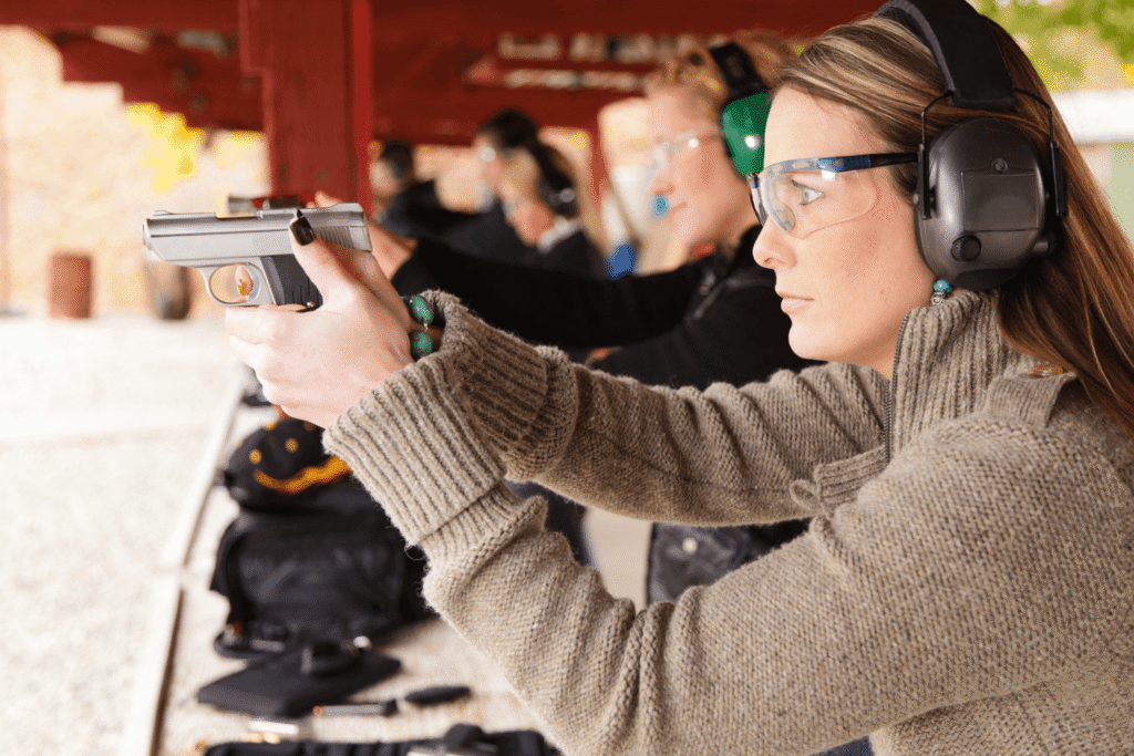Woman at an outdoor shooting range wearing protective gear and aiming her pistol towards a target