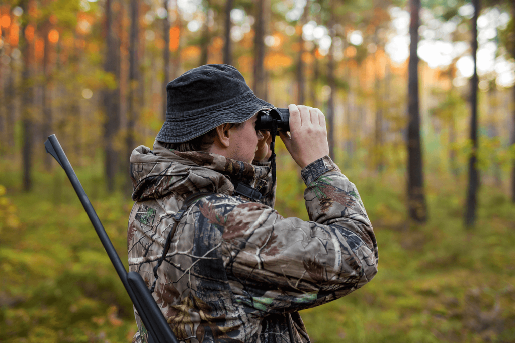 Man wearing camouflage sweater and black bucket hat looking through binoculars while hunting in the woods