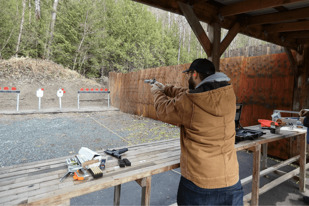 Man wearing a khaki jacket aiming his pistol at a target at an outdoor shooting range